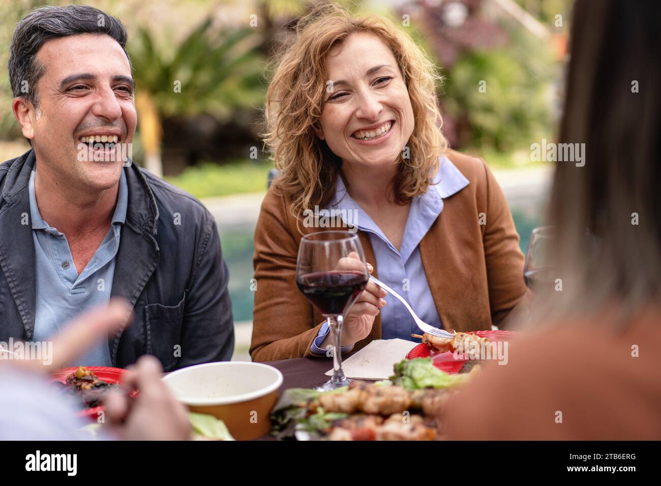 Zwei Freunde, die sich bei einem zwanglosen Essen im Freien einen Lachen und Wein teilen, umgeben von Natur und einer Vielzahl von Speisen. Stockfoto