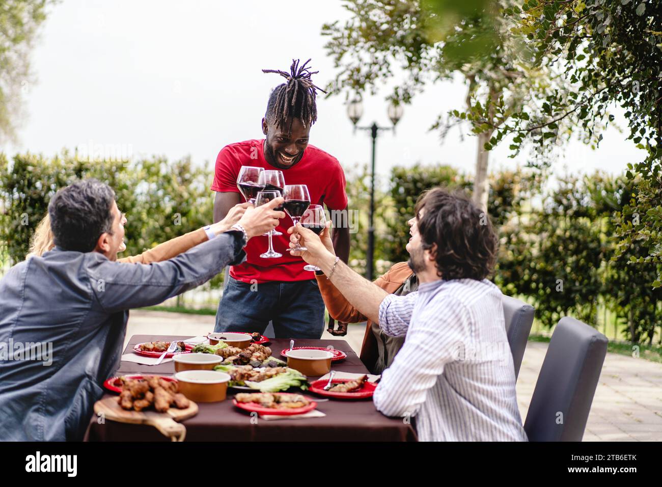 Verschiedene Freunde lachen und toasten mit Rotwein an einem Tisch im Freien, gefüllt mit köstlichem Essen. Stockfoto