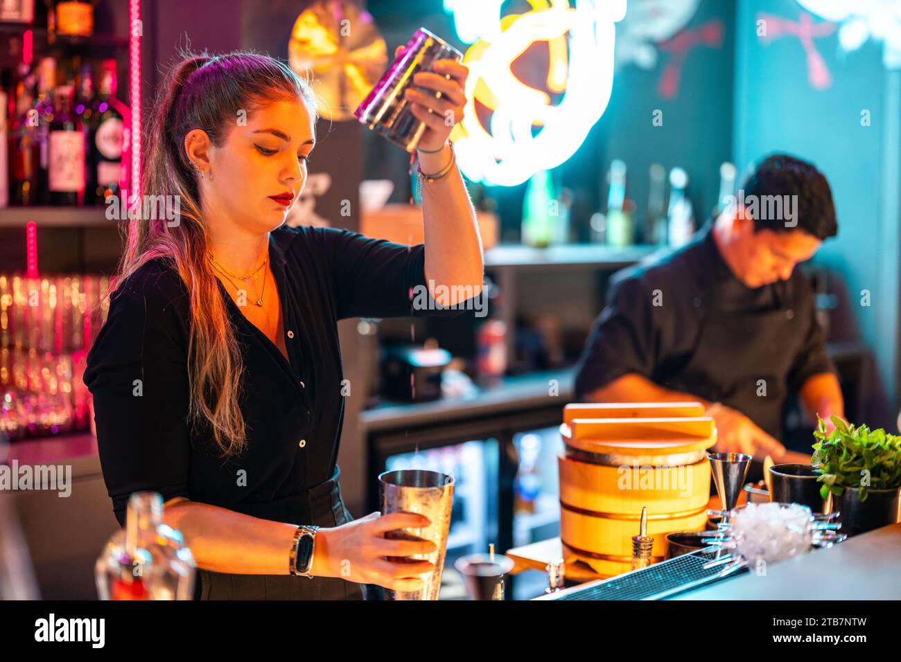 Seitenansicht der schönen jungen Barkeeper mit Konzentration, die alkoholische Getränke in Cocktailshaker an der Theke mischt, während sie in einem beleuchteten Nachtpub arbeiten Stockfoto