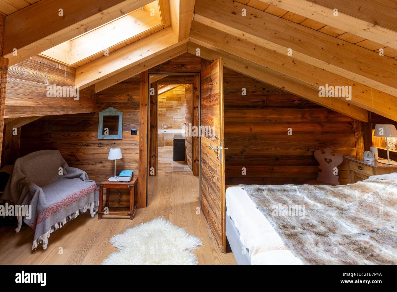 Ein warmes, rustikales Schlafzimmer im Dachgeschoss mit holzgetäfelten Wänden, Oberlicht und komfortablen Möbeln für eine gemütliche Atmosphäre in den Bergen. Stockfoto