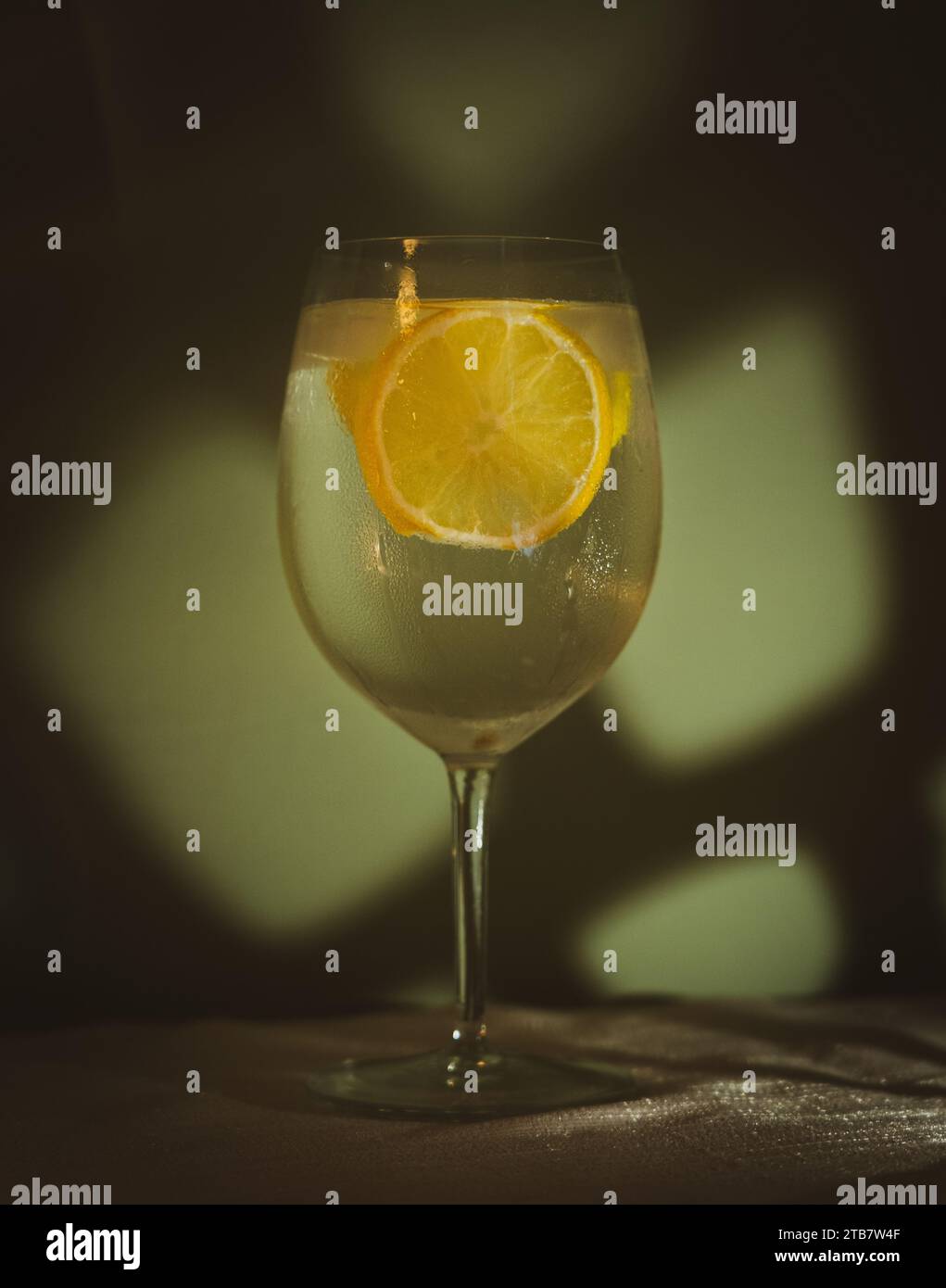 Gin-Tonic-Cocktail im Weinglas mit Eis- und Zitronenscheiben vor der hellgrünen Wand auf einem weißen Tischtuch Stockfoto