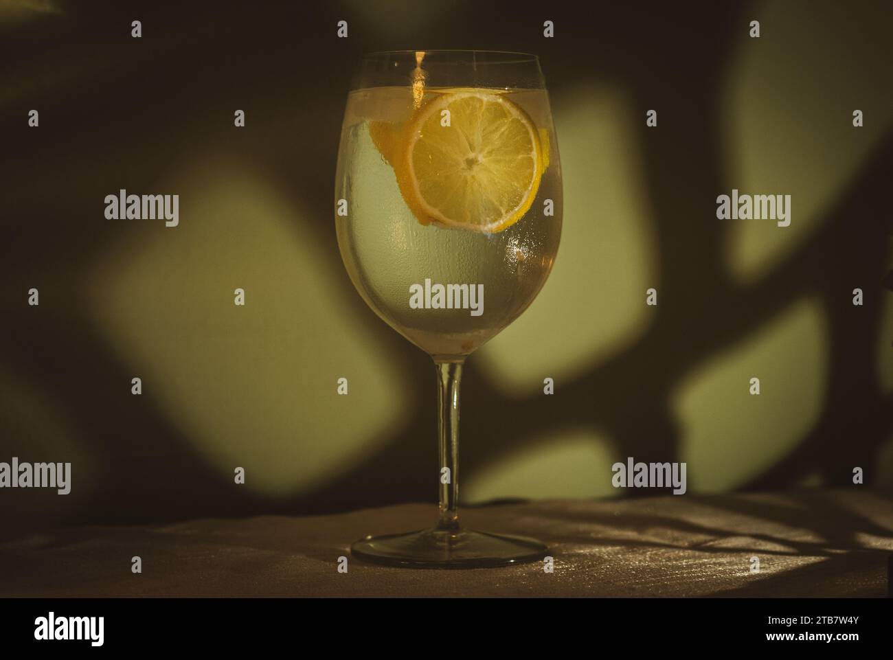 Gin-Tonic-Cocktail im Weinglas mit Eis- und Zitronenscheiben vor der hellgrünen Wand auf einem weißen Tischtuch im Licht Stockfoto