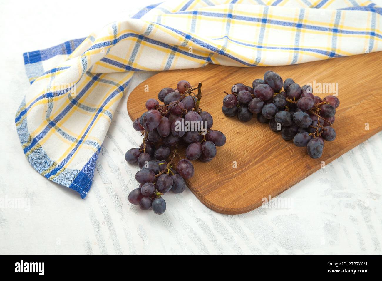 Schneidebrett mit einem großen Haufen reifer dunkelblauer Trauben auf weiß. Lila Weintrauben mit blauem Küchentuch auf weißem hölzernem Hintergrund. Stockfoto