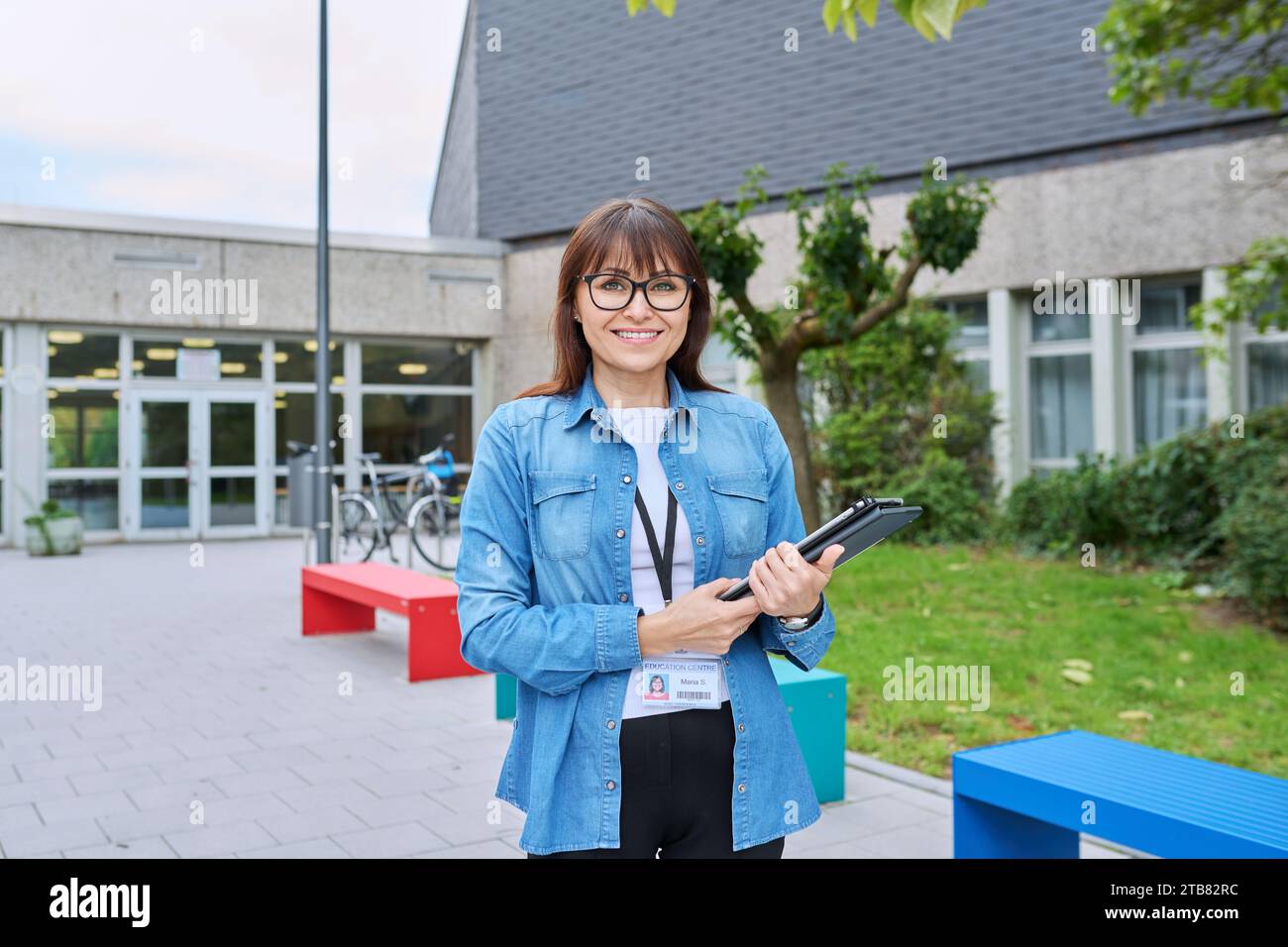 Schullehrerin mittleren Alters mit digitalem Tablet posiert in der Nähe des Schulgebäudes Stockfoto