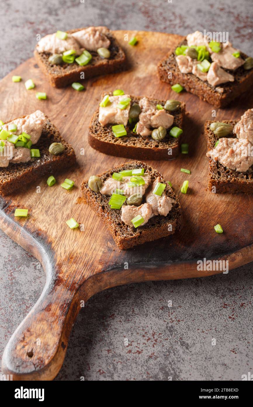 Roggentoast mit Kabeljauleber, grünen Zwiebeln und Kapern in Nahaufnahme auf einem Holzbrett auf dem Tisch. Vertikal Stockfoto