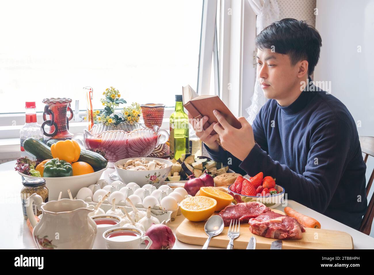 Junger asiatischer Mann, der ein Buch liest und alleine am Esstisch speisen und im Winter an Thanksgiving Essen und Trinken feiern kann Stockfoto