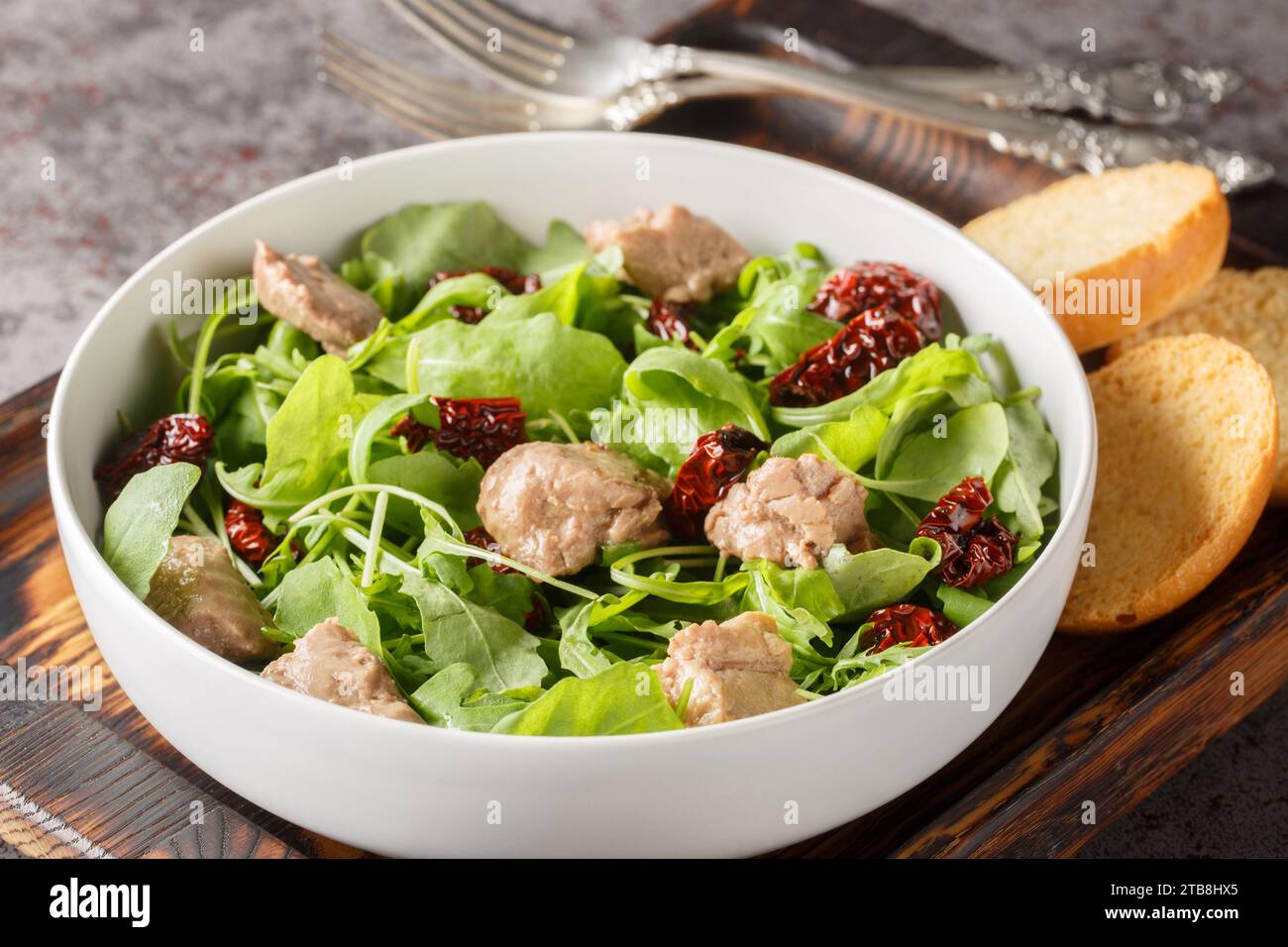 Köstlicher Kabeljaulebersalat mit Rucola und sonnengetrockneten Tomaten, serviert mit Toast in einer Schüssel auf dem Tisch. Horizontal Stockfoto