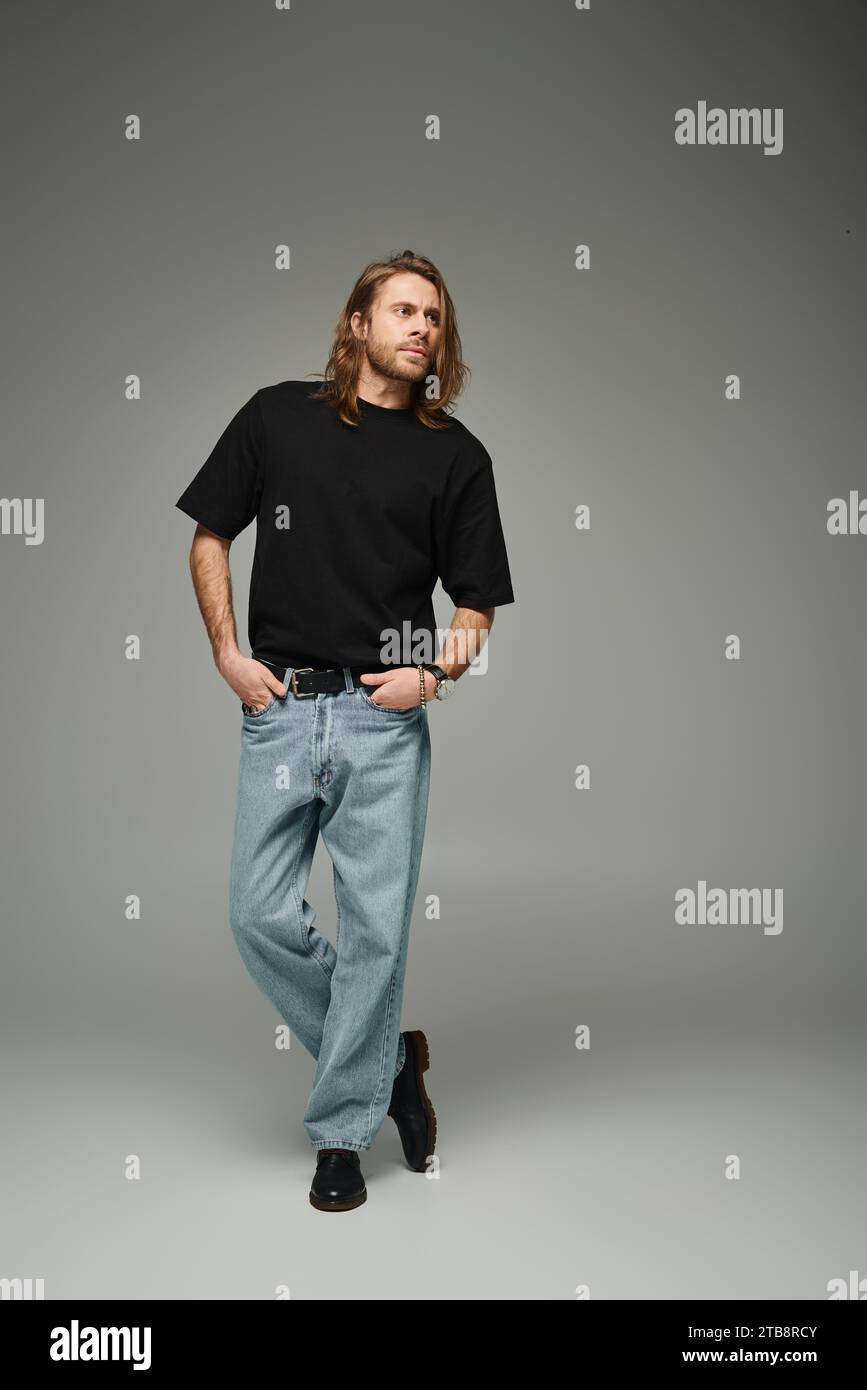 Die ganze Länge eines bärtigen, gutaussehenden Mannes in schwarzem T-Shirt und Jeans, der mit der Hand in den Taschen auf Grau steht Stockfoto