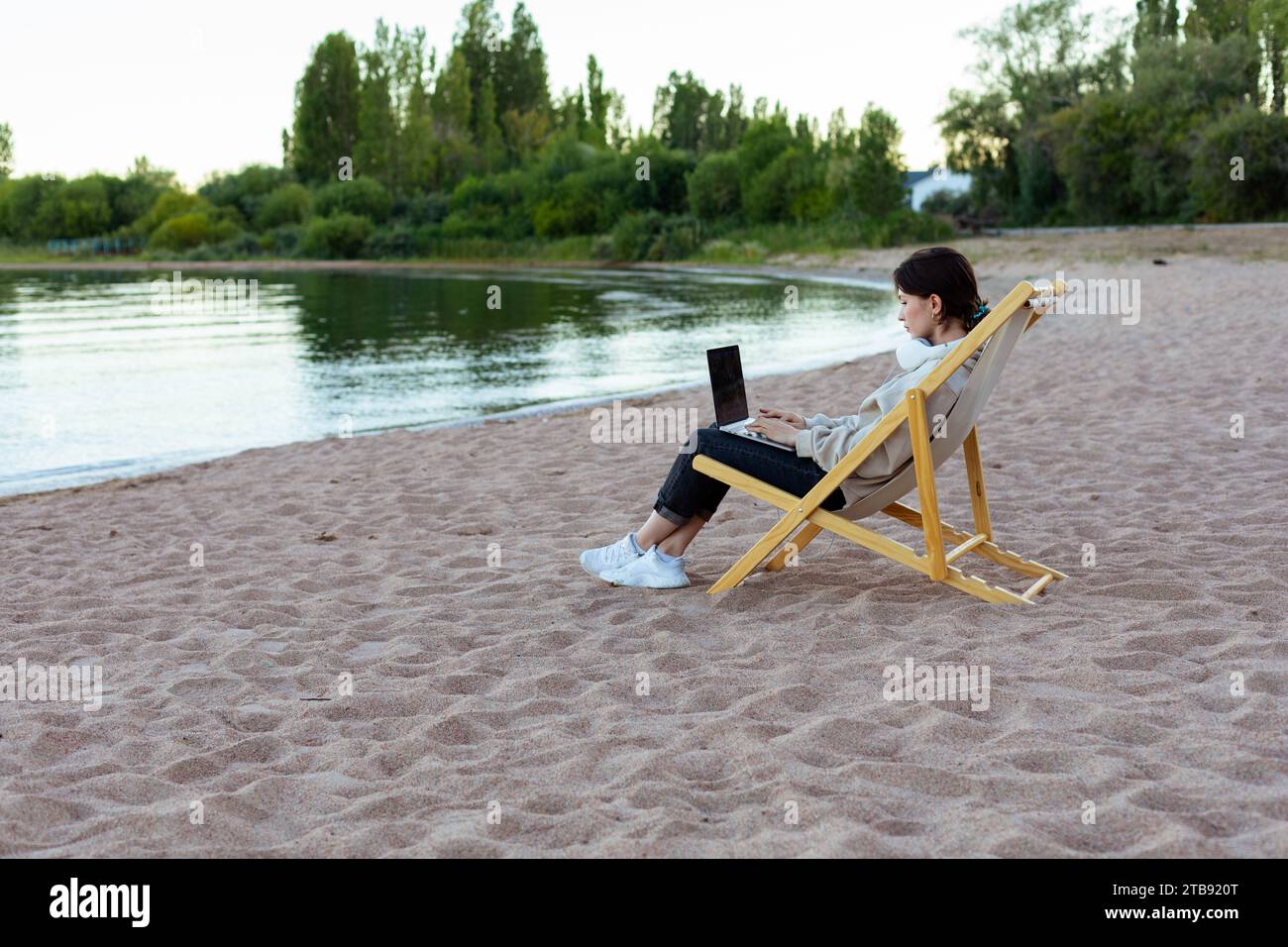 Freischaffende Frau in Kopfhörern, die seitlich am Laptop am See sitzt Stockfoto