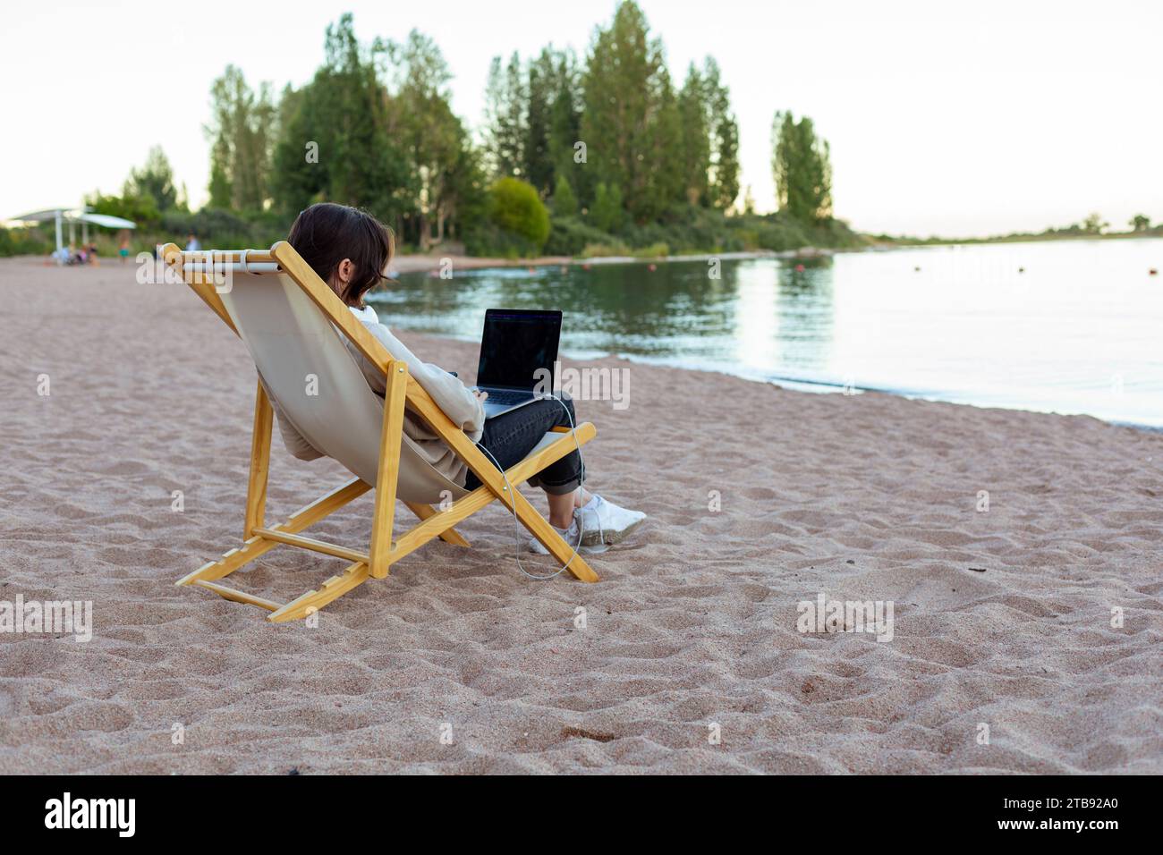 Freischaffende Frau in Kopfhörern, die seitlich am Laptop am See sitzt Stockfoto