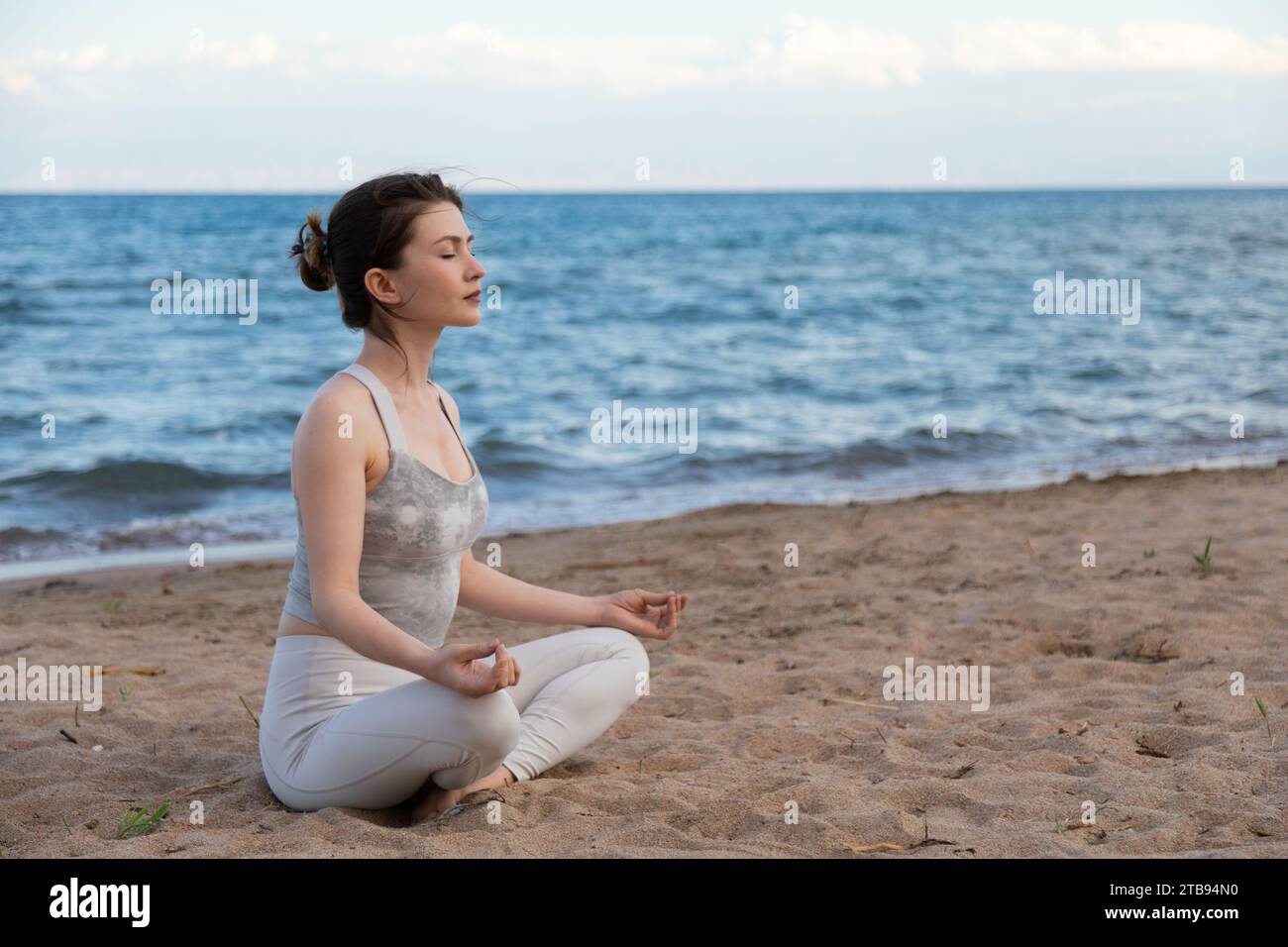 Junge schöne Frau, die Yoga im Freien praktiziert, sitzt am Seeufer in Lotusposition, entspannt, tief atmet und meditiert. Auf Sand in der Nähe von W sitzen Stockfoto