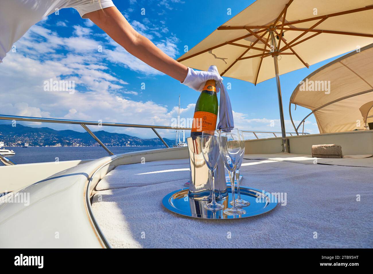 Hände mit weißen Handschuhen, die ein Glas Veuve Clicquot Champagner an Bord einer Charteryacht mit Deck und blauem Meer dahinter gießen. Stockfoto