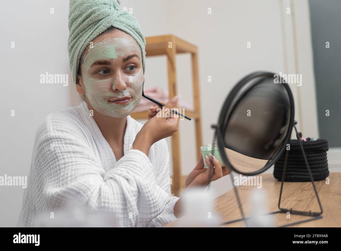 Die Frau trägt eine Tonmaske auf, blickt in den Spiegel und gönnt sich zu Hause ihrer organischen Gesichtsbehandlung. Stockfoto