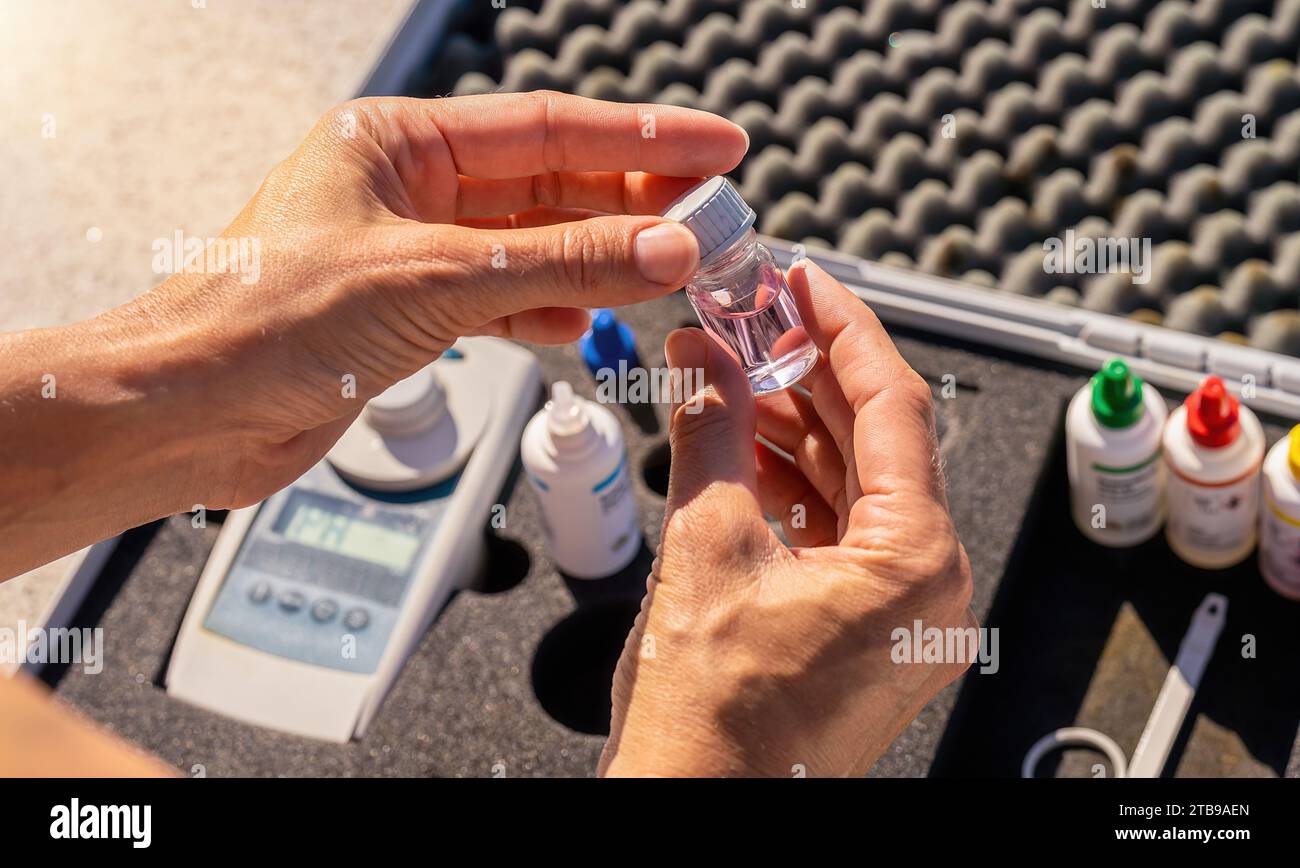 Hände, die eine kleine leere Testflasche mit farbcodierten Reagenzflaschen und digitalem Gerät in einem Gehäuse im Hintergrund öffnen Stockfoto