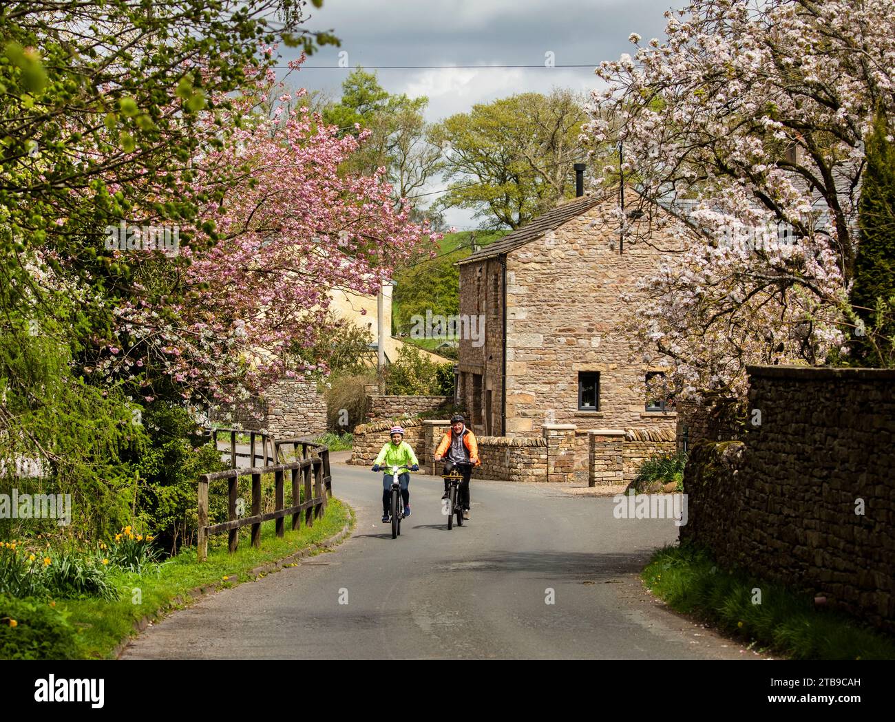 Mann und Frau Radfahrer fahren E-Bikes in einem hübschen Dorf im Frühling, Hartley. Kirkby Stephen, North Pennines, Cumbria Stockfoto