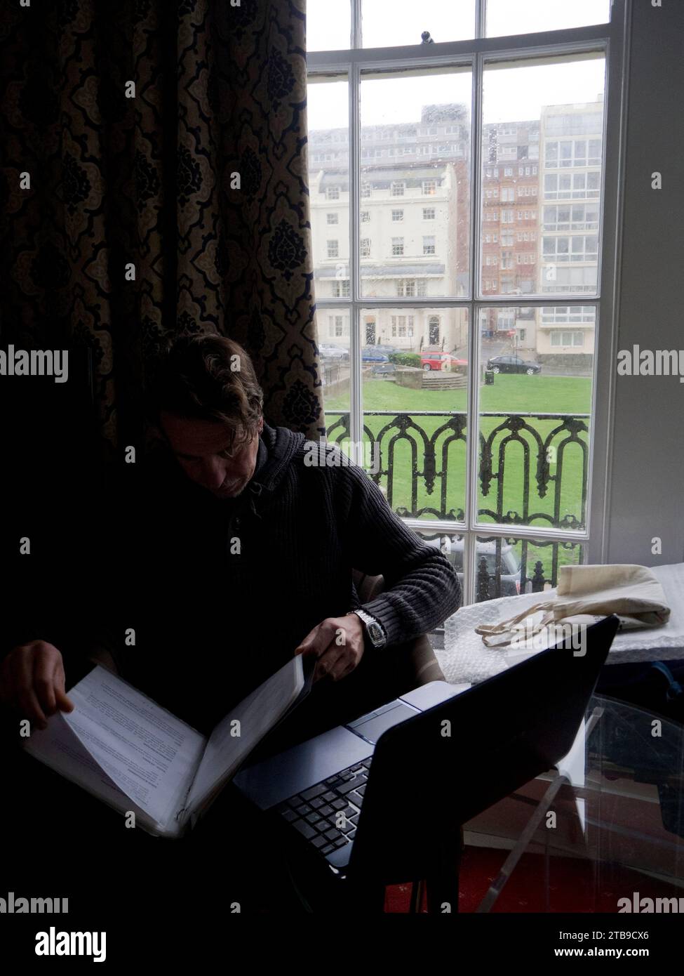 Mann, der an einem regnerischen Tag in einem Hotelzimmer in Brighton mit Laptop am Fenster sitzt und Dokumente liest; Brighton, East Sussex, England, Großbritannien Stockfoto
