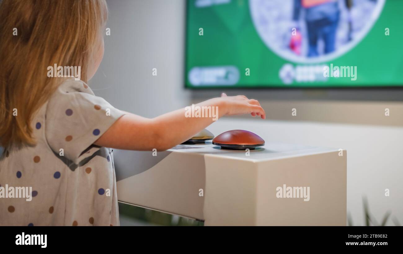 Ein kleines Mädchen, das auf einen großen Fernsehbildschirm mit interaktiven Inhalten blickt und ein Wissensspiel spielt. Stockfoto