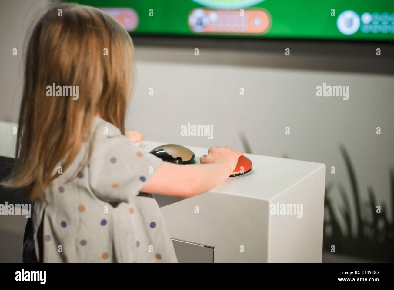 Ein kleines Mädchen, das auf einen großen Fernsehbildschirm mit interaktiven Inhalten blickt und ein Wissensspiel spielt. Stockfoto