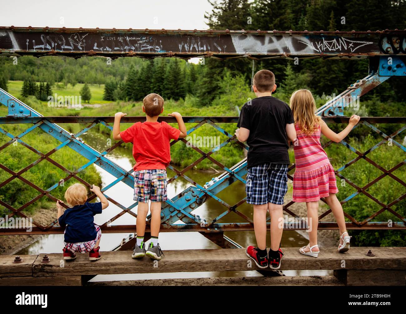 Blick von hinten auf vier Geschwister mit Blick auf ein Metallgeländer zusammen von einer Brücke über einen Fluss auf einem Naturlehrpfad Stockfoto