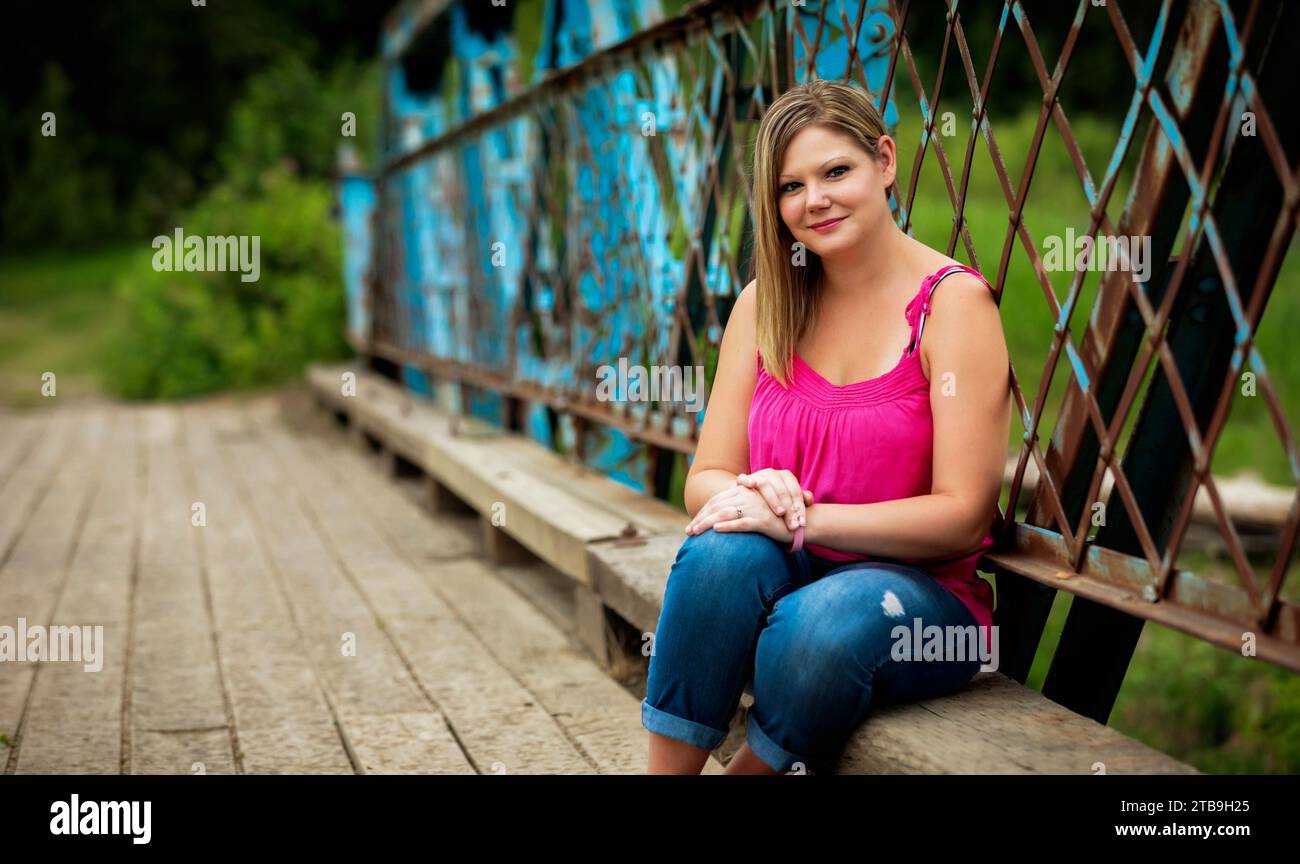 Nahaufnahme eines Porträts einer Frau, die auf einer Holzbank entlang einer Brücke sitzt, während eines Naturspaziergangs in einem Park, posiert für die Kamera Stockfoto