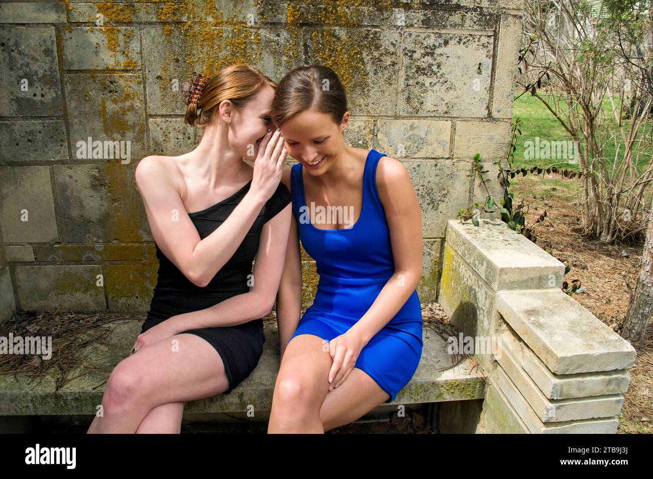 Zwei junge Frauen in Minikleidern sitzen auf einer Bank in einem Gartenbereich und flüstern einander Geheimnisse zu; Lincoln, Nebraska, USA Stockfoto