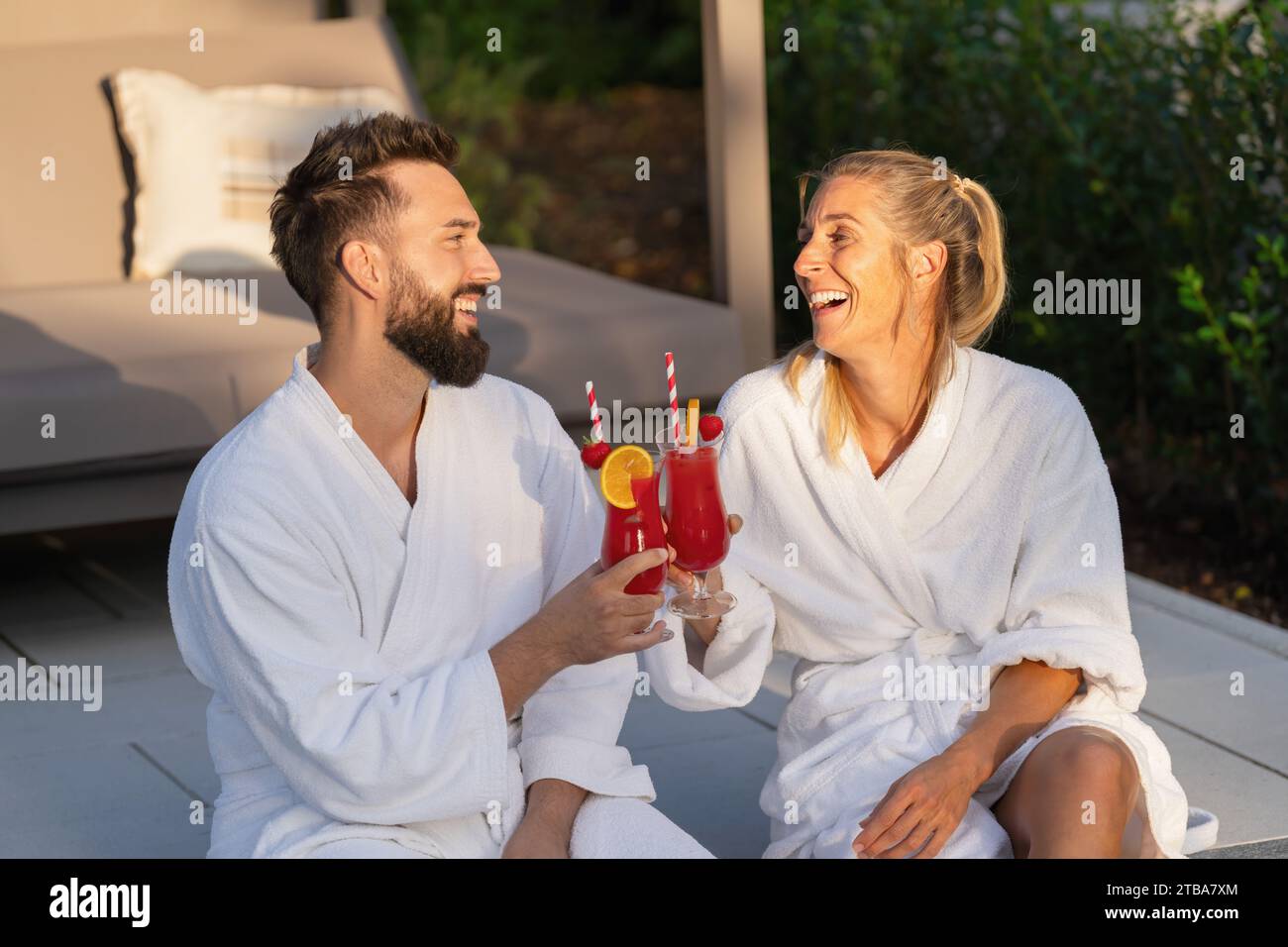 Fröhliches Paar in weißen Bademänteln, das Gläser mit roten Getränken anklickt, draußen zur goldenen Stunde in einem Hotel Stockfoto