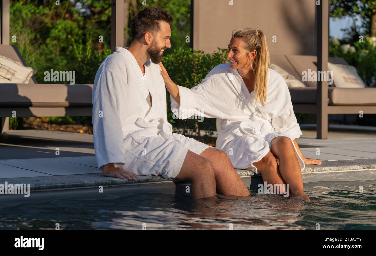 Paare in weißen Bademänteln sitzen mit Füßen im Pool und genießen einen sonnigen Tag im Spa Wellness Hotel Stockfoto