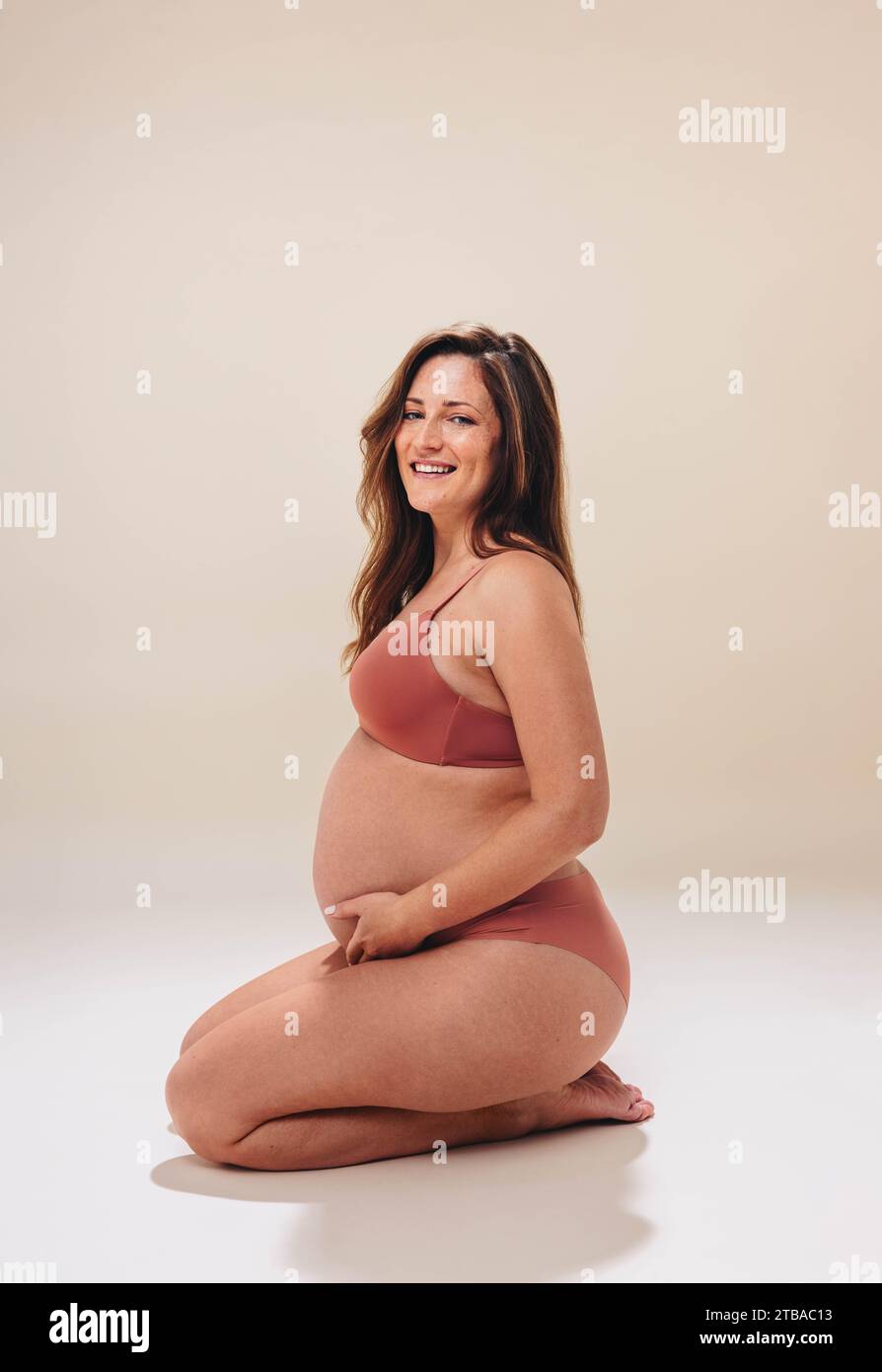 Glückliche zukünftige Mutter kniet in einem Studio auf dem Boden, hält ihr wachsendes Baby und lächelt in die Kamera. Schwangere kaukasische Frau zeigt Aufregung Stockfoto