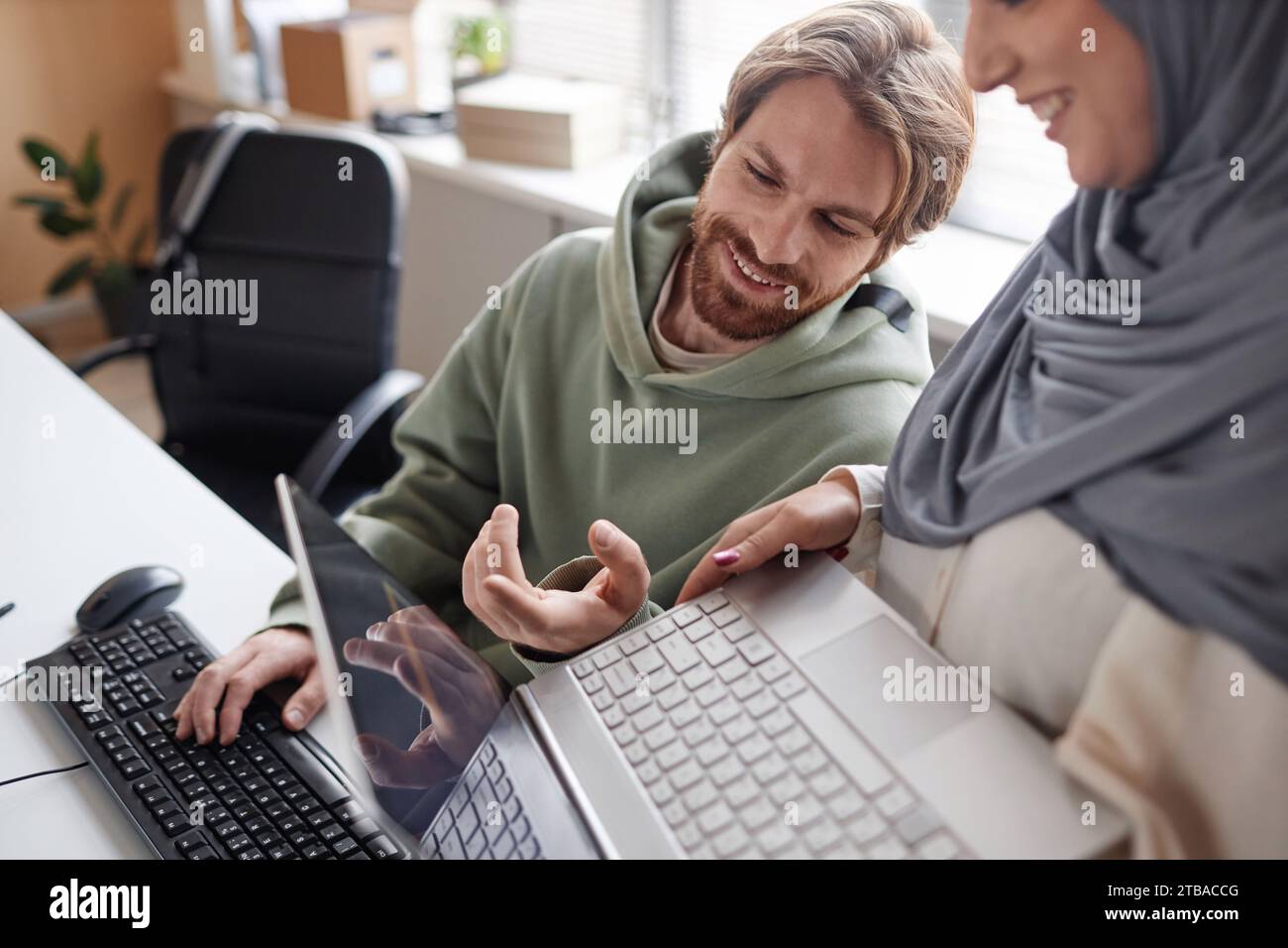 Porträt eines bärtigen jungen Mannes, der mit einem muslimischen Kollegen im IT-Büro spricht und lächelt, während er auf den Laptop zeigt Stockfoto