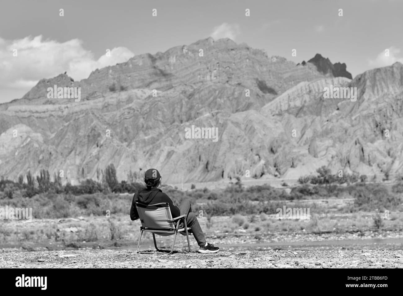 Rückansicht eines asiatischen Mannes, der im Stuhl auf dem Flussbett sitzt und auf die Klippen der Schlucht in Xinjiang, China, blickt, schwarz-weiß Stockfoto