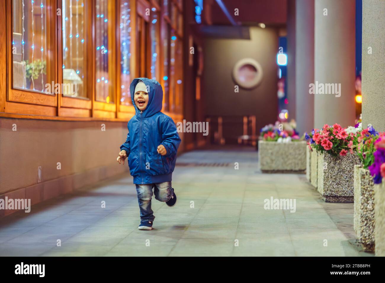 Happy Little Boy läuft entlang der Galerie zwischen Säulen mit Blumen und Gebäudefassade am späten Abend Stockfoto