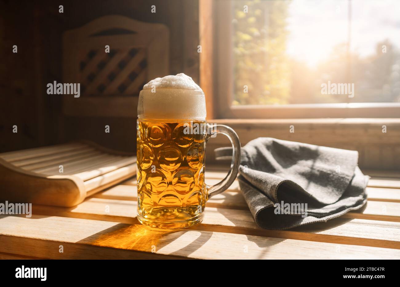 Bierkrug mit geschäumtem Bier ruhen auf einem Felsvorsprung in einer Sauna und fangen Sonnenlicht ein. In der Nähe befinden sich finnische Saunahüte auf einer Holzbank. Bild des Spa- und Wellnesskonzepts Stockfoto