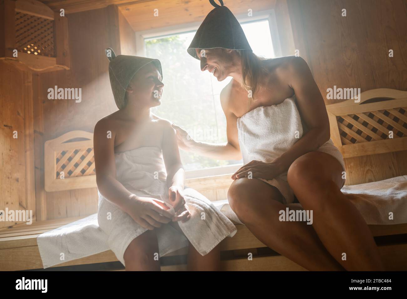 Mutter und Kind lächeln einander in einer finnischen Sauna an, beide tragen Filzhüte Stockfoto