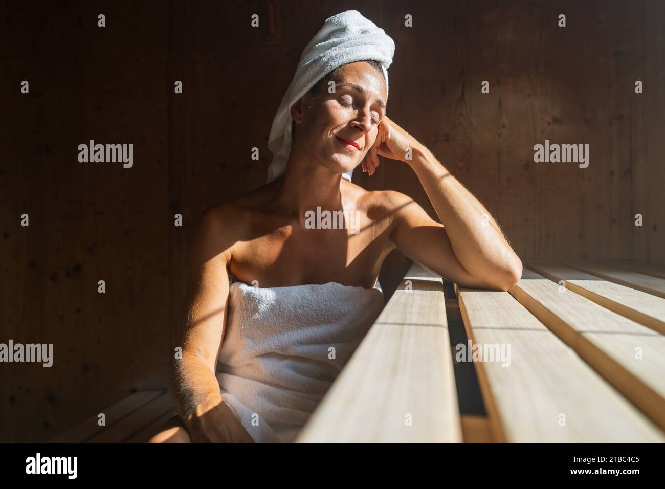 Frau in einer finnischen Sauna, die sich zurücklehnt, die Augen geschlossen, mit einem Handtuch um den Kopf gewickelt und in einem Spa-Resort sonnig beleuchtet Stockfoto