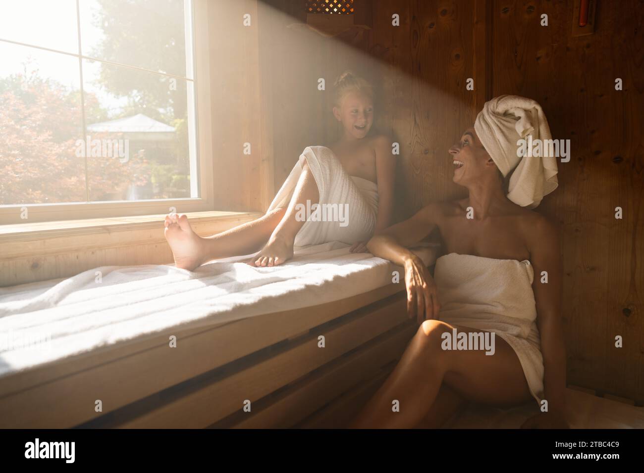 Mutter und Tochter in der Sauna, Mutter mit einem Handtuch auf dem Kopf, beide lächelnd und sitzend Stockfoto