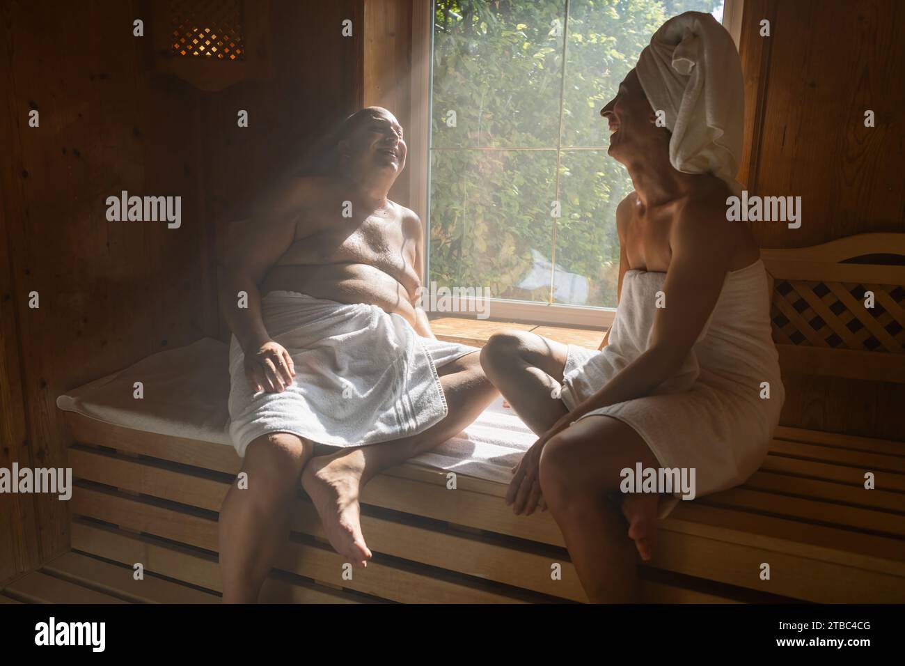 Mann und Frau in einer finnischen Sauna, lachend, Frau mit einem Handtuch auf dem Kopf, beide sitzen im Wellnesshotel Stockfoto