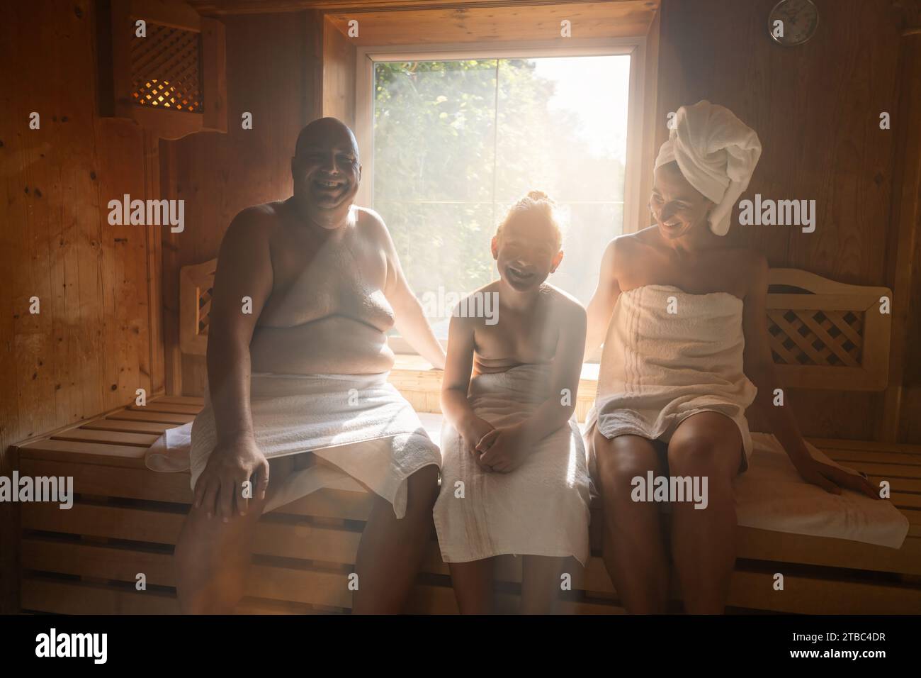 Familie in einer finnischen Sauna lacht zusammen, Mann links, Frau mit Handtuch auf dem Kopf rechts, Kind mitten im Wellnesshotel Stockfoto