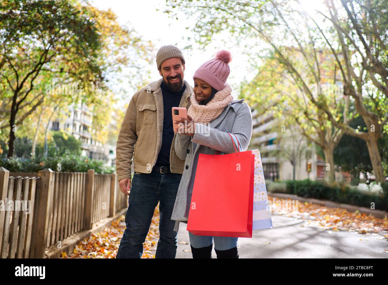 Junge interrassische Paare, die das Telefon benutzen, während sie auf der Straße spazieren gehen und gemeinsam weihnachtseinkäufe machen. Stockfoto