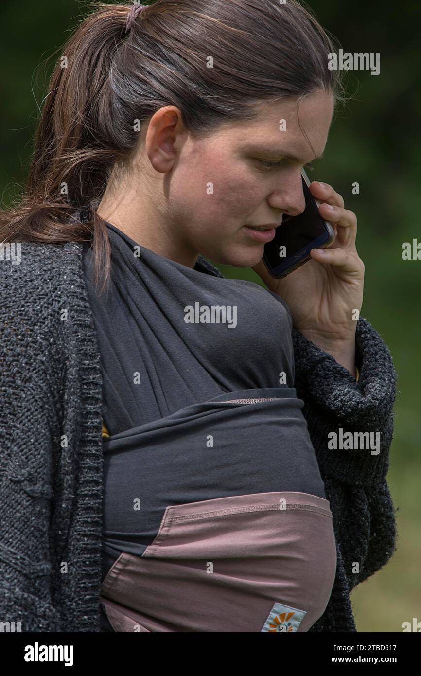 Junge Mutter am Telefon mit ihrem Smartphone, ihr Baby in eine Babytragetasche gewickelt, Mecklenburg-Vorpommern, Deutschland Stockfoto
