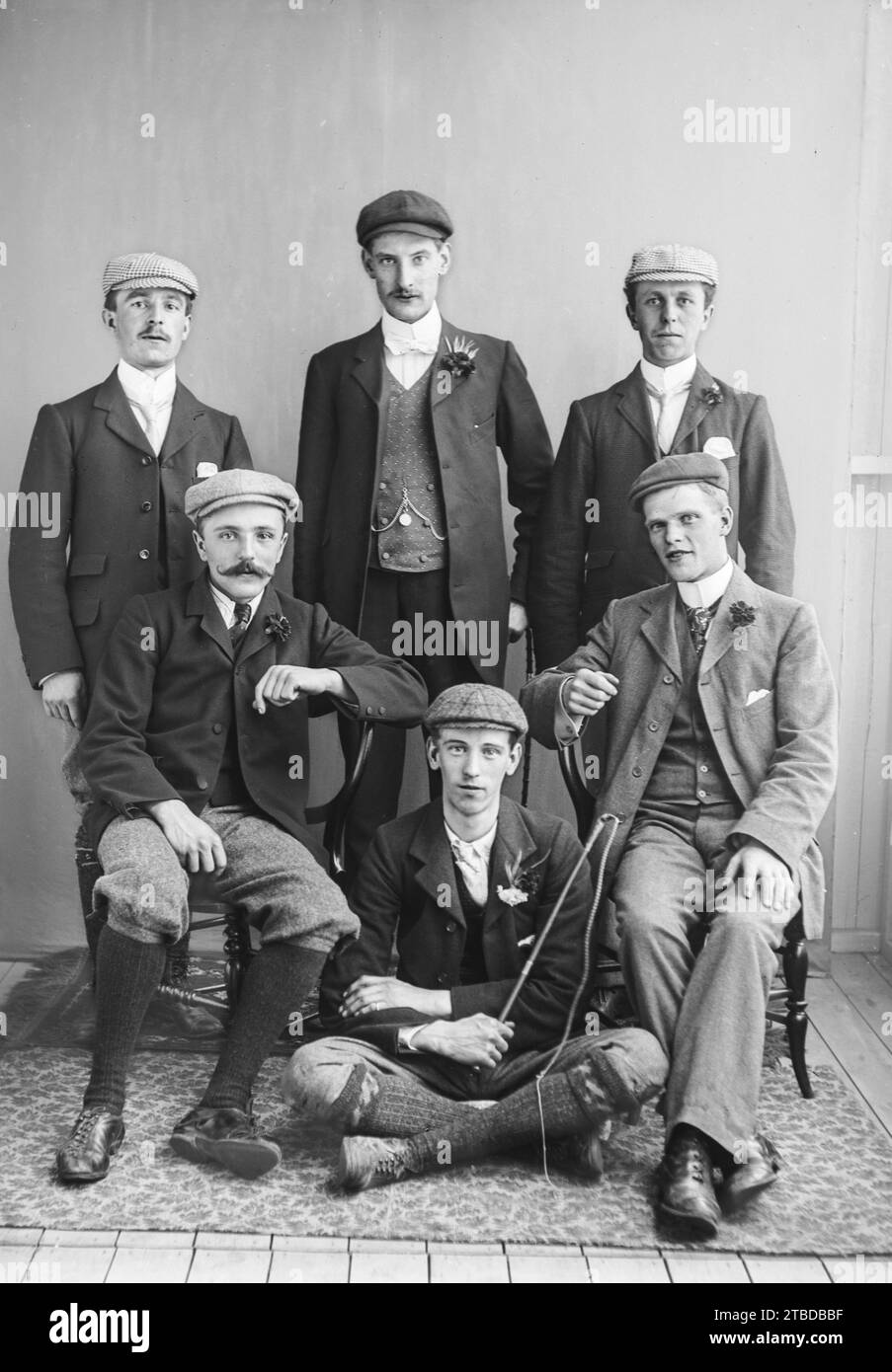 Edwardianisches Studio-Porträt von sechs Männern. Drei stehen und zwei sitzen auf Stühlen und eines mit überkreuzten Beinen auf dem Boden. Knopflöcher, lange Socken, Peitsche, Kappen. Von einer alten Glasplatte negativ. C1900 – 1910 Stockfoto