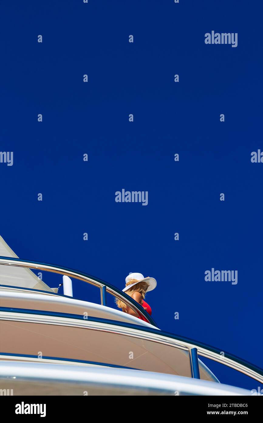 Mädchen mit rotem Kleid und Strohhut, das auf dem Seitendeck einer Superyacht steht und sich an der Edelstahlschiene mit tiefblauem Himmel festhält. Stockfoto