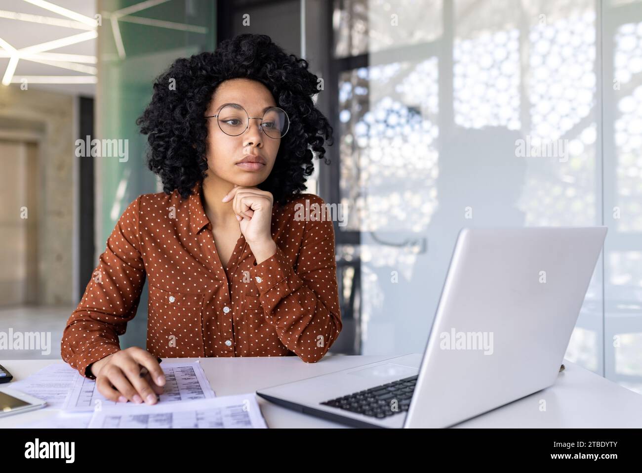 Nachdenkliche Afroamerikanerin in Brille, die im Büro arbeitet, am Schreibtisch vor dem Laptop sitzt, ihr Kinn mit der Hand hält, auf den Bildschirm blickt und nachdenkt. Stockfoto