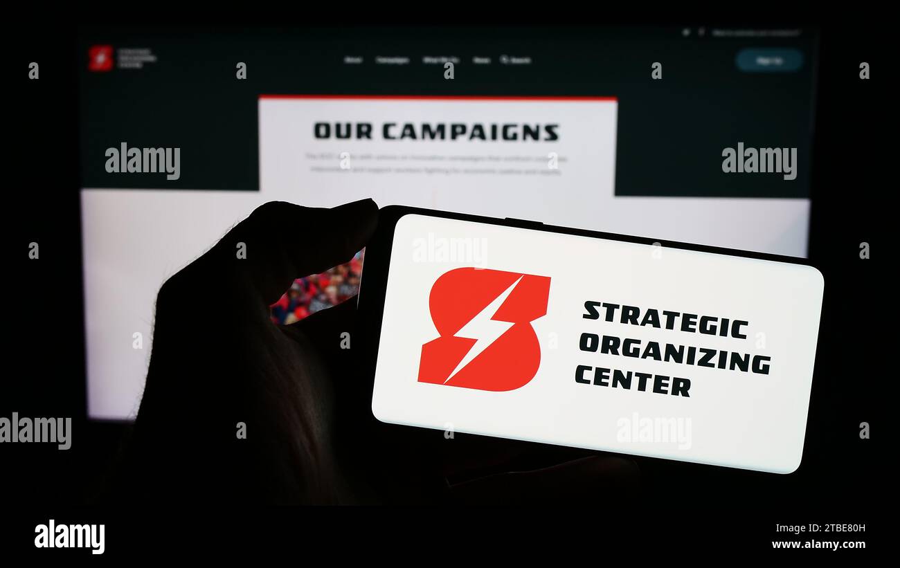 Person, die ein Mobiltelefon mit dem Logo der gewerkschaftskoalition Strategic Organizing Center (SOC) vor der Webseite hält. Schwerpunkt auf dem Display des Telefons. Stockfoto