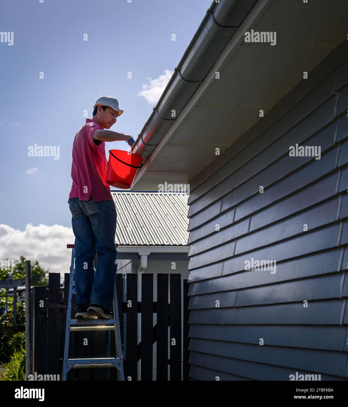 Mann, der auf der Leiter steht und die Rinne reinigt. Hauswartungsarbeiten. Vertikales Format. Auckland. Stockfoto