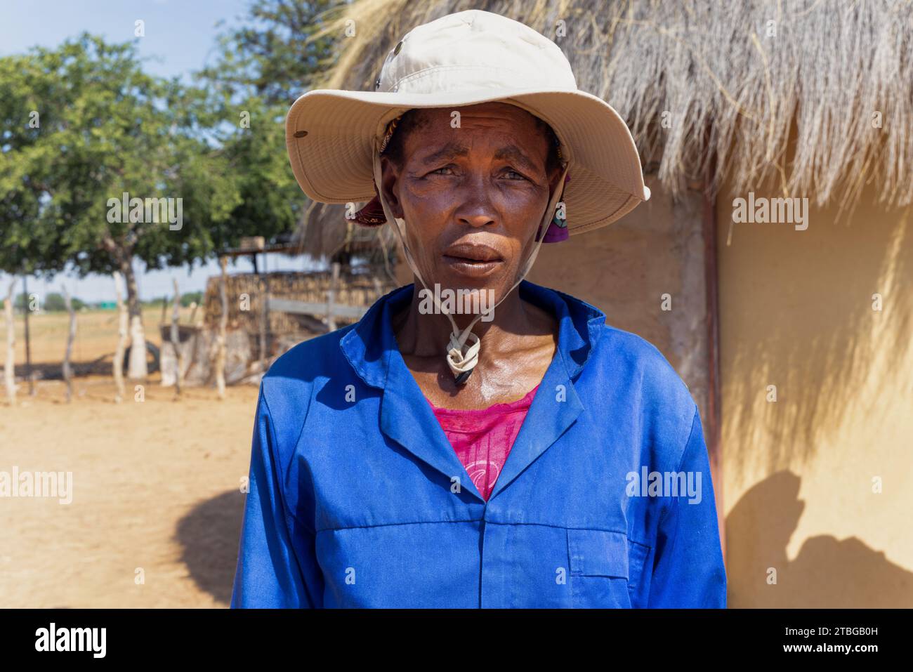 afrikanisches Dorfleben, Frau in Arbeitskleidung, die einen Hut vor ihrem strohgedeckten Schlammhaus im Hof trägt Stockfoto