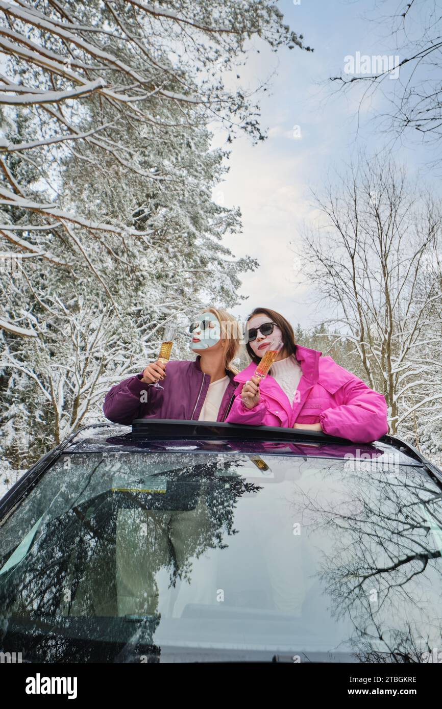 Zwei lustige Frauen mit kosmetischen Masken im Gesicht, die ein Glas Champagner aus der Autoluke halten Stockfoto