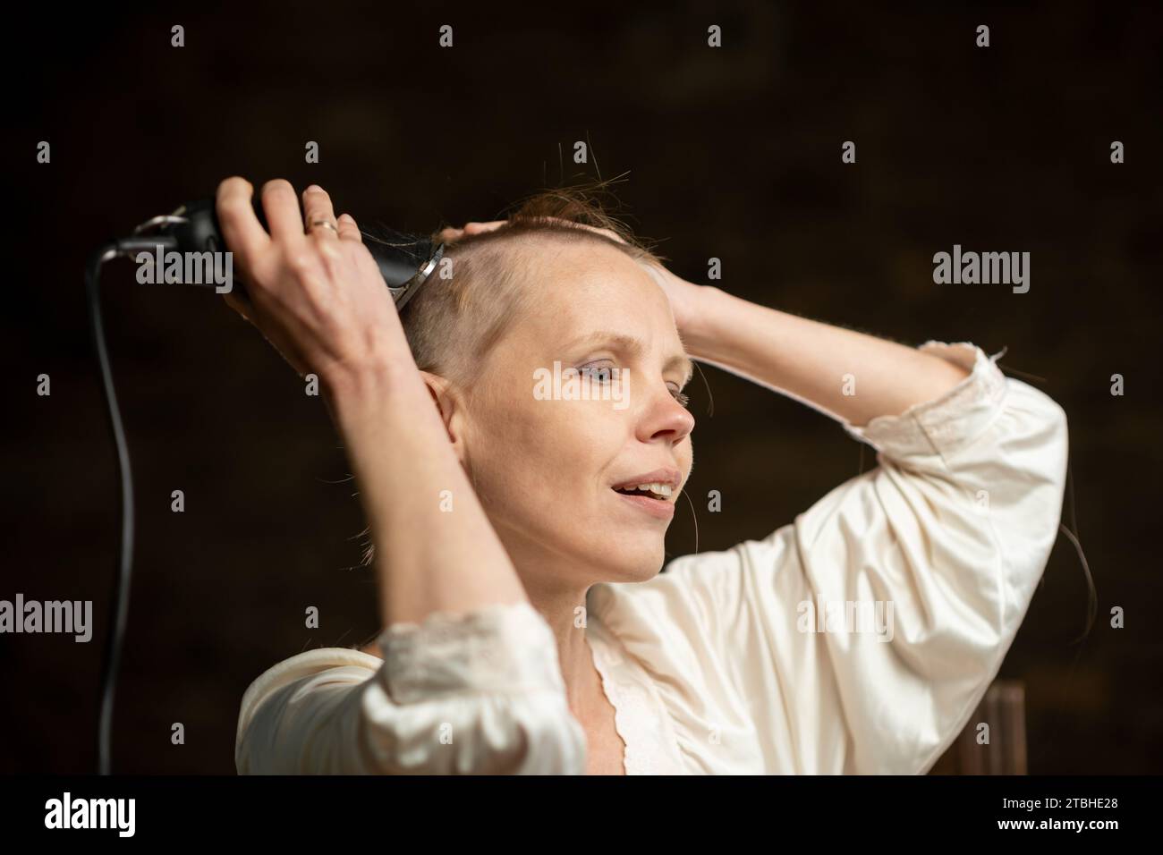 Eine Frau nimmt sich mit einem Haarschneider ihr Haar. Kahle Frau mittleren Alters in einem weißen Hemd auf dunklem Hintergrund. Stockfoto