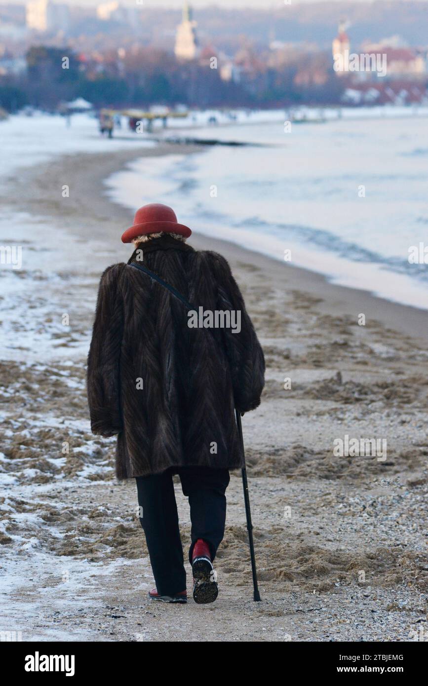 Elegant gekleidete einsame Seniorin mit einem Stock, die an einem kalten Wintertag am schneebedeckten Danzig-Strand entlang in Richtung der Kurstadt Sopot an der Ostseeküste, Polen, Europa, EU Stockfoto
