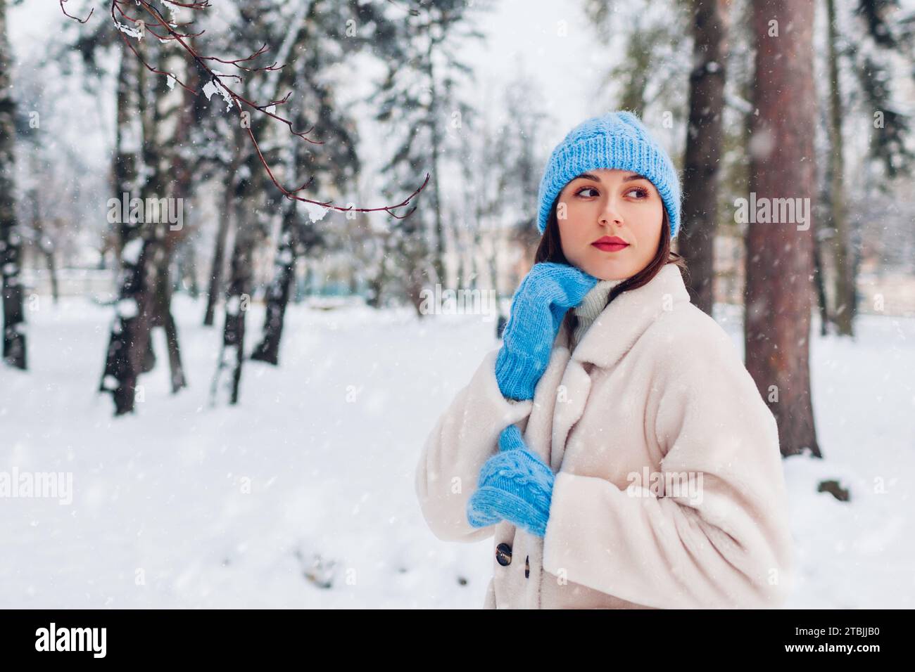 Nahaufnahme Porträt einer jungen Frau, die im Winterpark unter Schnee läuft, mit weißem Pelzmantel, blauem Strickmütze und Fäustlingen. Stilvolle, gemütliche warme Kleidung Stockfoto
