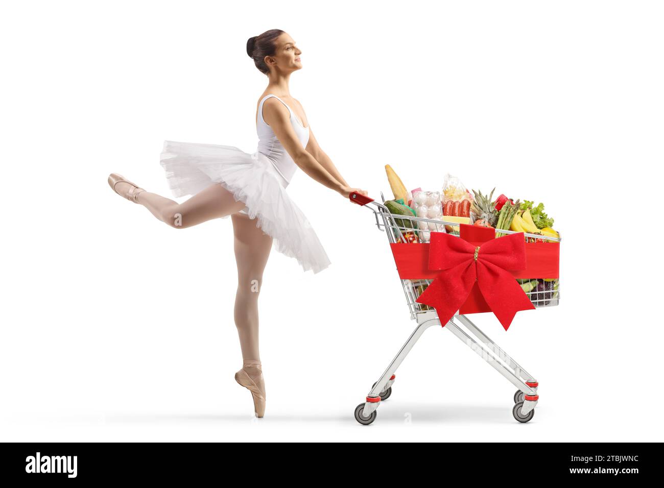 Profilaufnahme einer Ballerina in voller Länge, die tanzt und einen Einkaufswagen mit Essen und roter Schleife auf weißem Hintergrund schiebt Stockfoto
